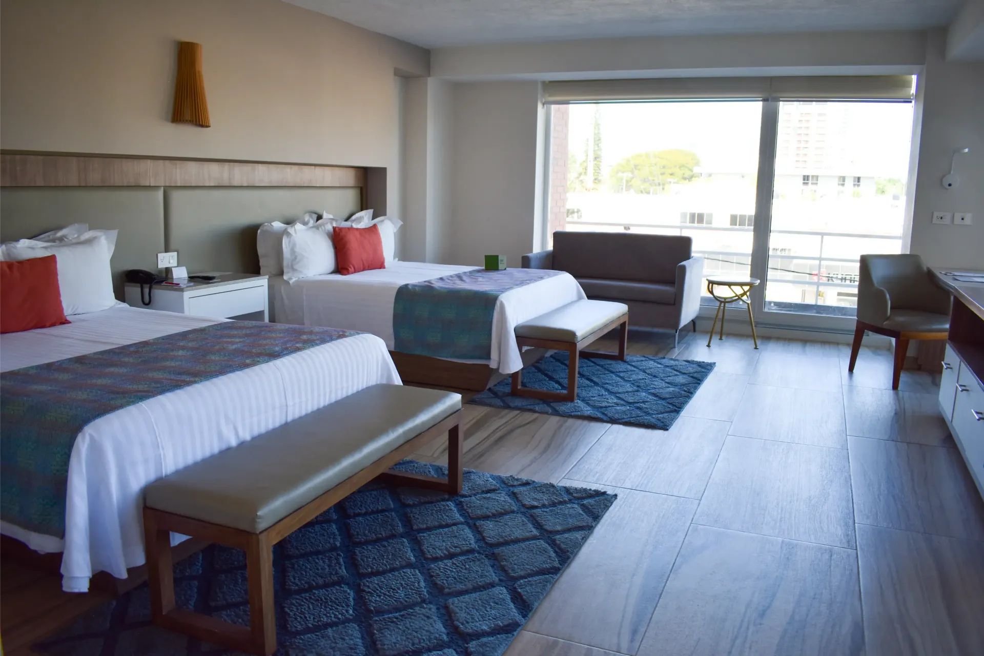 Habitación de hotel con dos camas individuales, bancos y una gran ventana con vistas a un balcón.