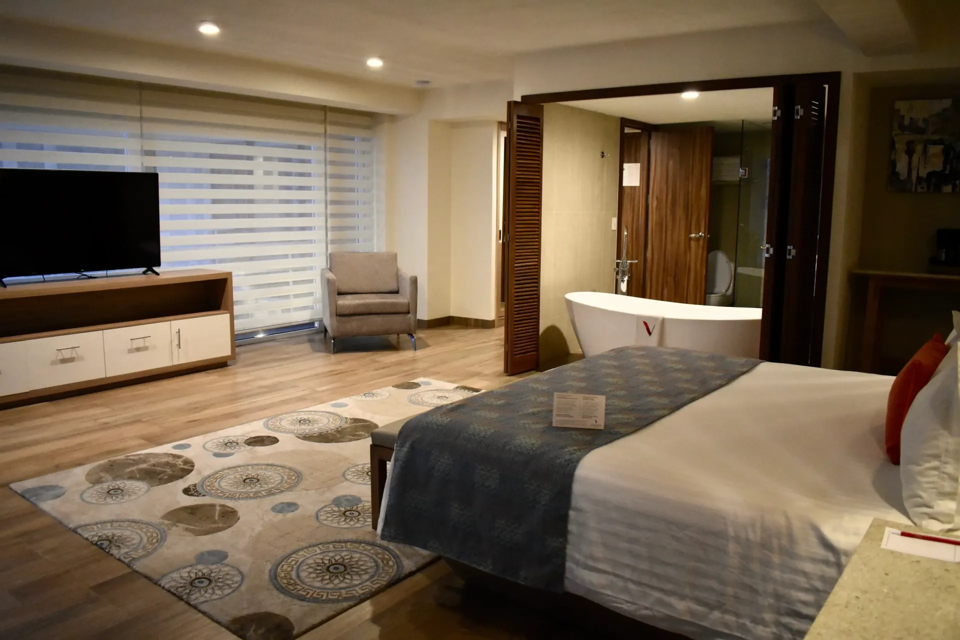 Amplia habitación de hotel con cama king, TV, sillón, alfombra y baño visible a través de las puertas abiertas.