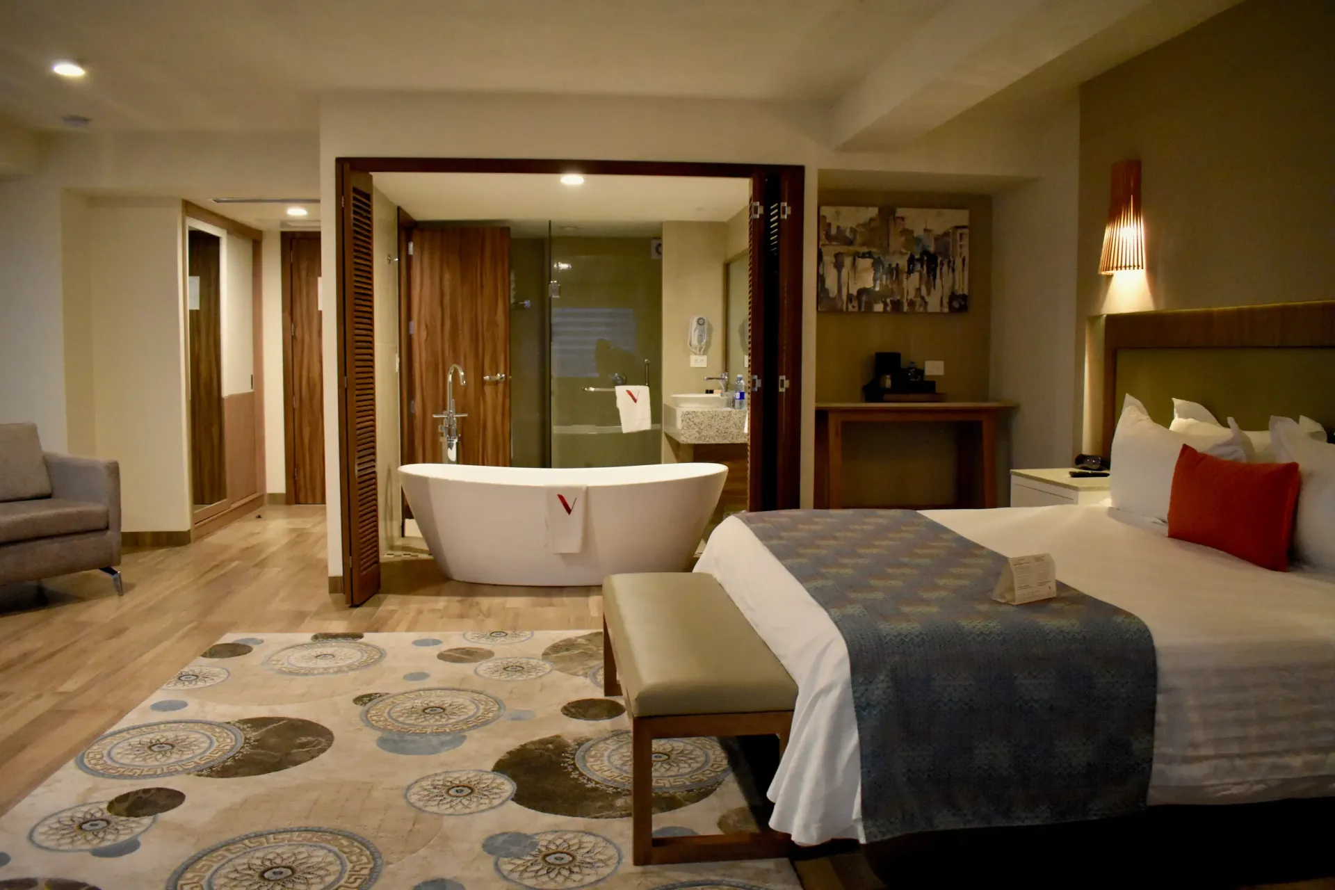 Habitación de hotel moderna con cama king, bañera independiente, sofá e iluminación cálida.
