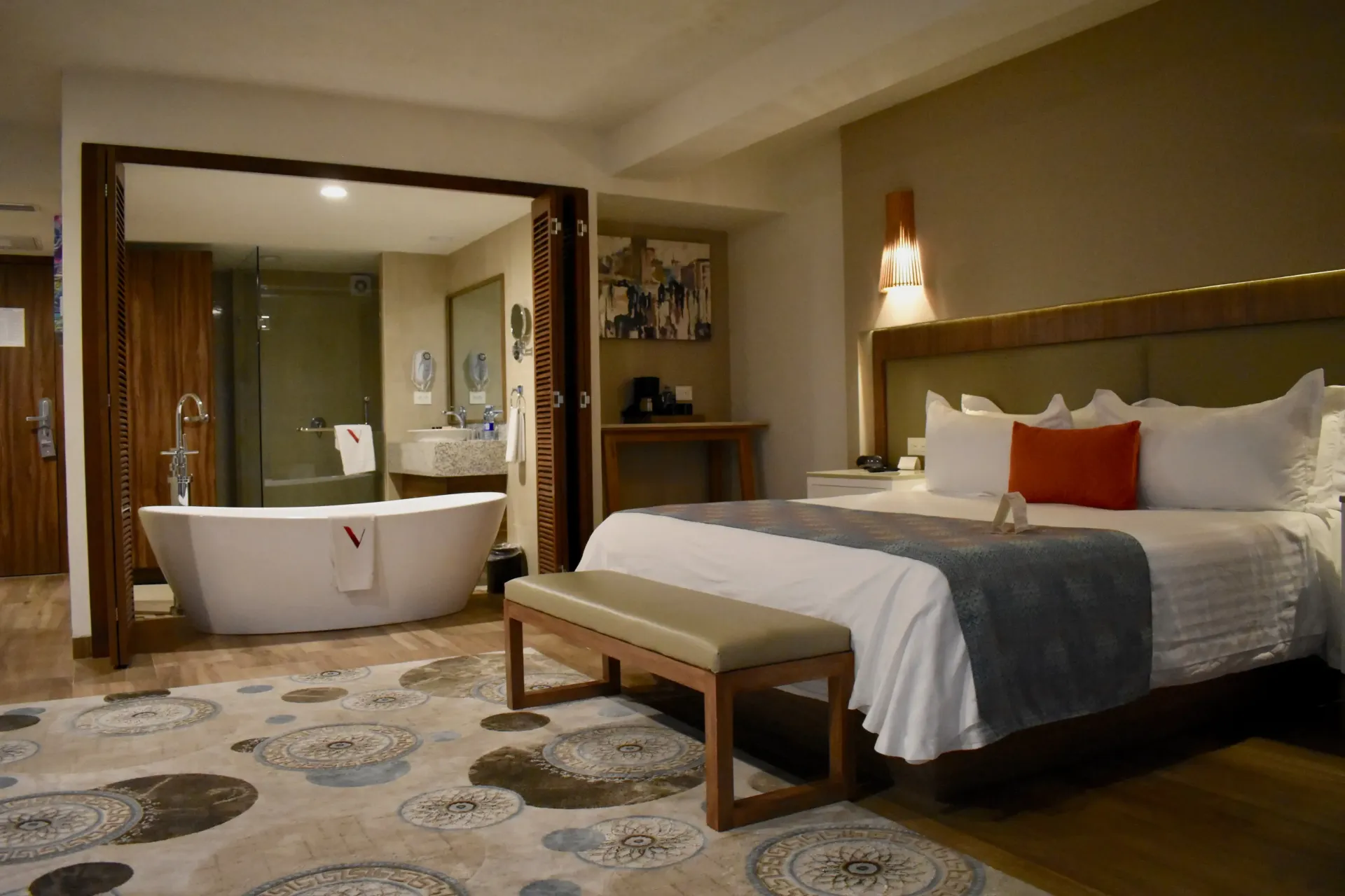 Habitación de hotel moderna con cama king size, banco y baño abierto con bañera independiente.