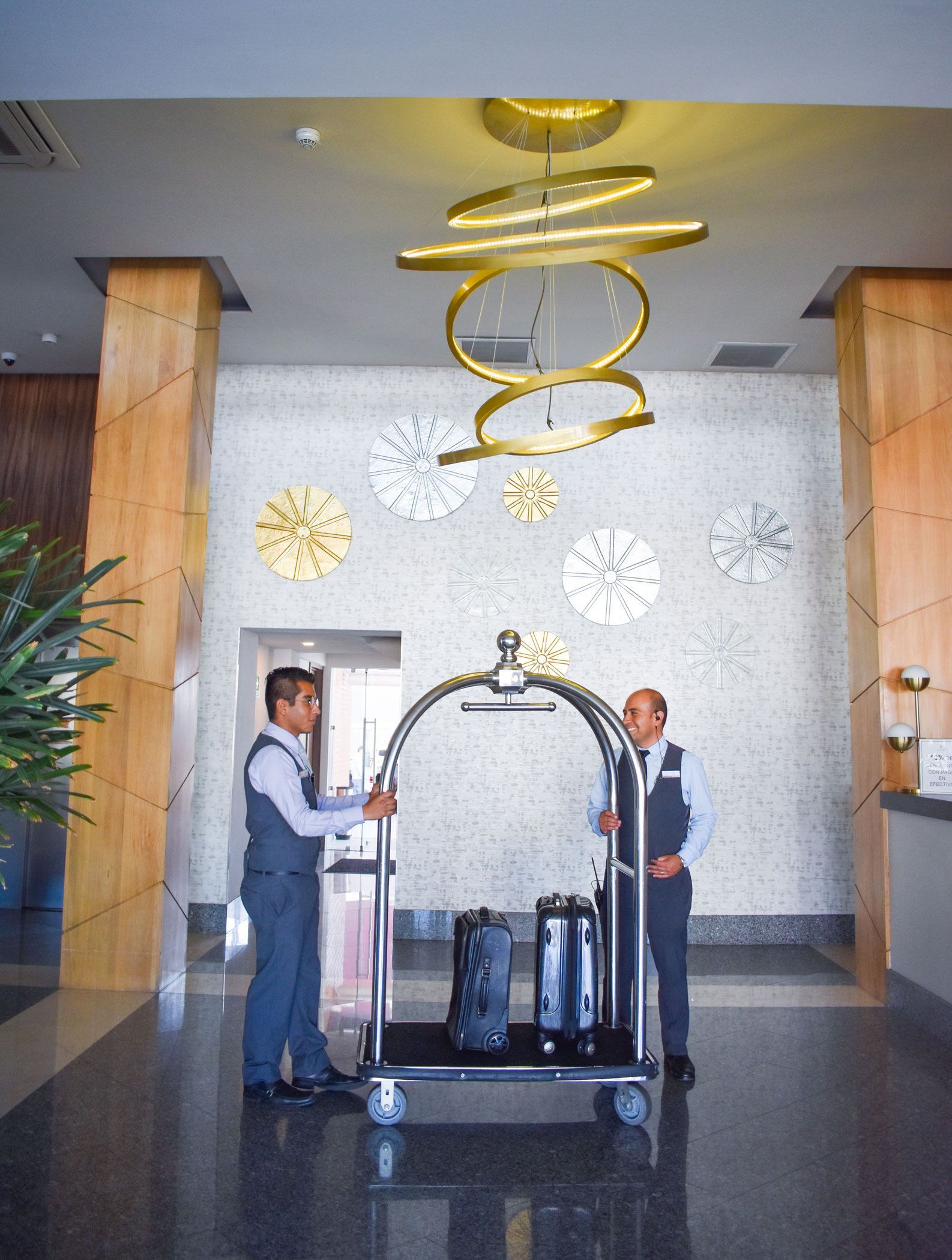 Vestíbulo del hotel con dos botones sosteniendo un carrito de equipaje bajo una decoración colgante de anillos dorados.