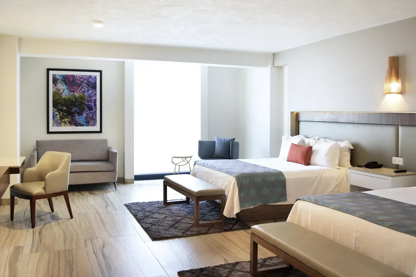 Habitación de hotel moderna con dos camas, sillas de color beige, obras de arte y una ventana luminosa.