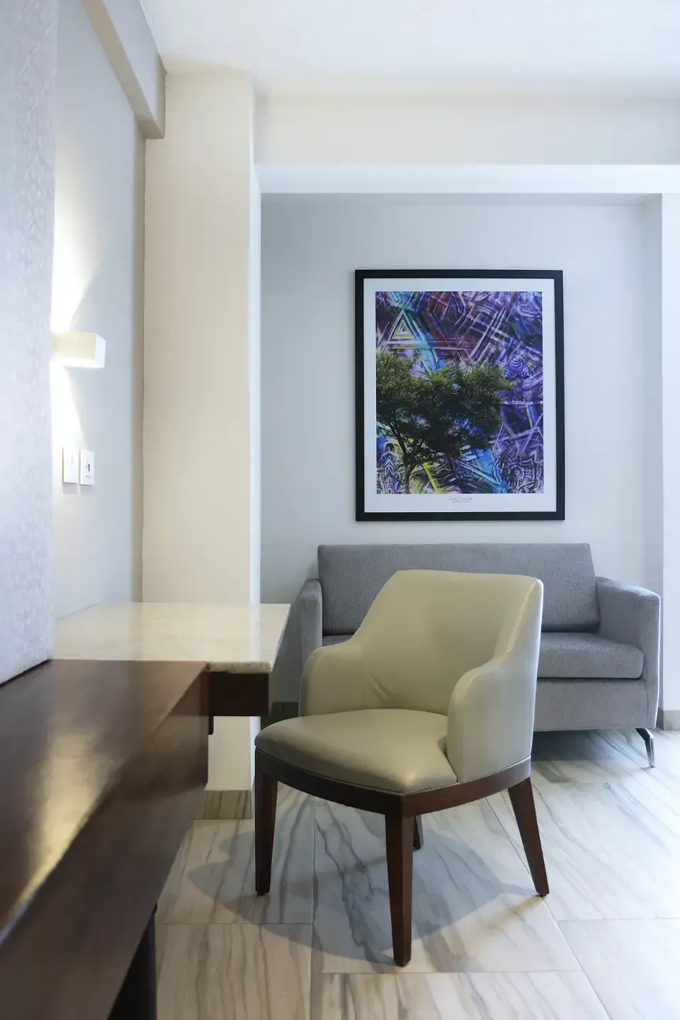 Sala de estar moderna con silla beige, sofá gris y obra de arte abstracta enmarcada.