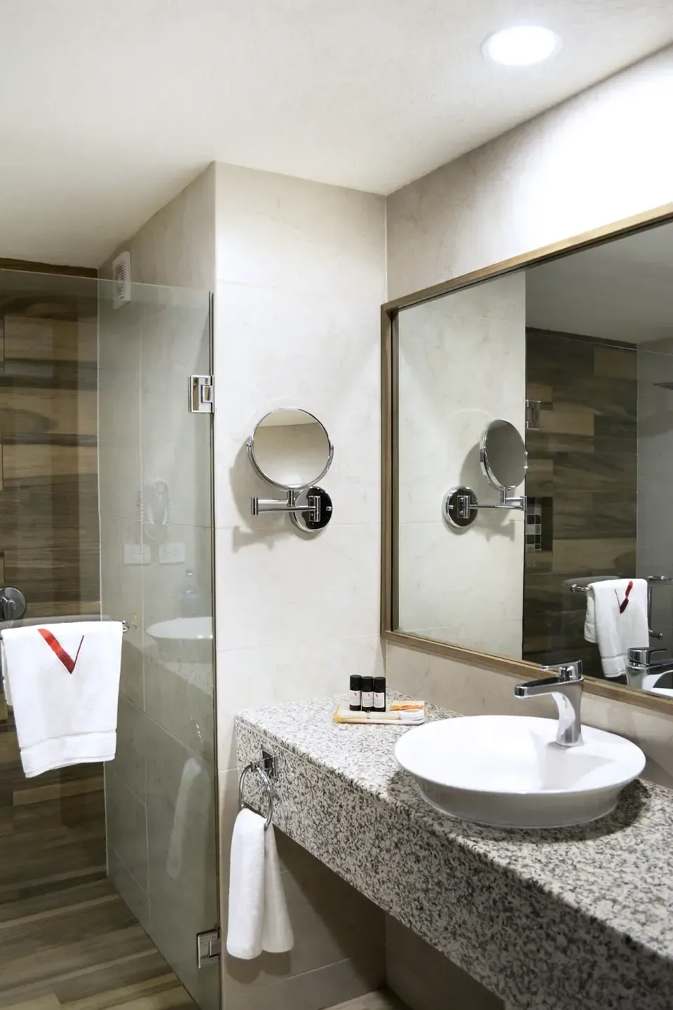 Baño de hotel moderno con ducha de cristal, tocador de granito, espejo redondo y lavabo blanco.