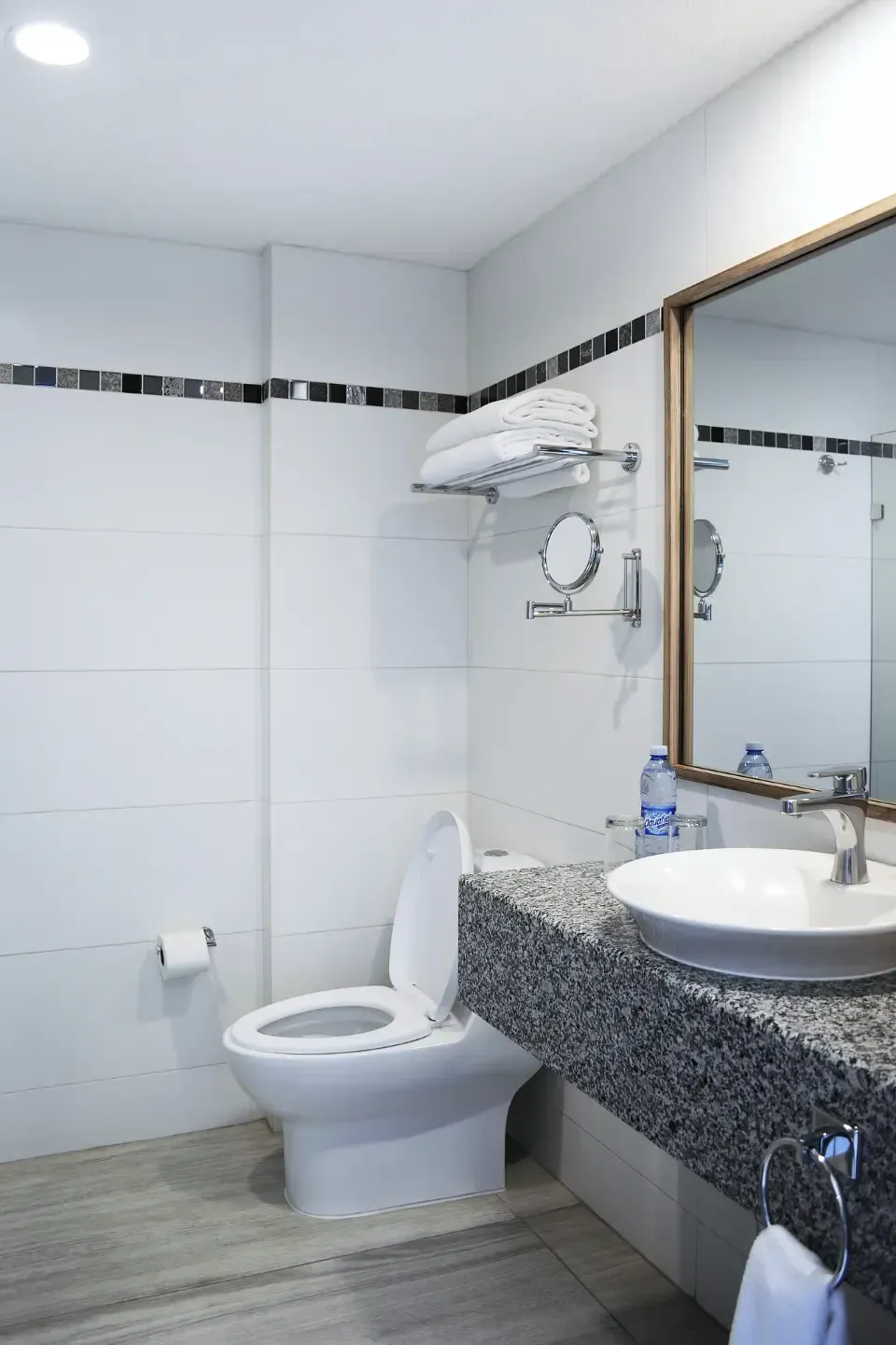 Baño moderno con azulejos blancos, inodoro, lavabo de granito, espejo grande y toallero.