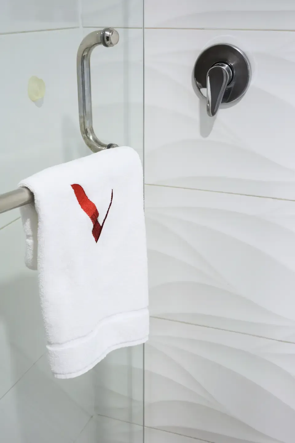 Toalla blanca con bordado de pájaro rojo colgada en una cabina de ducha de cristal con barra y grifo cromados.