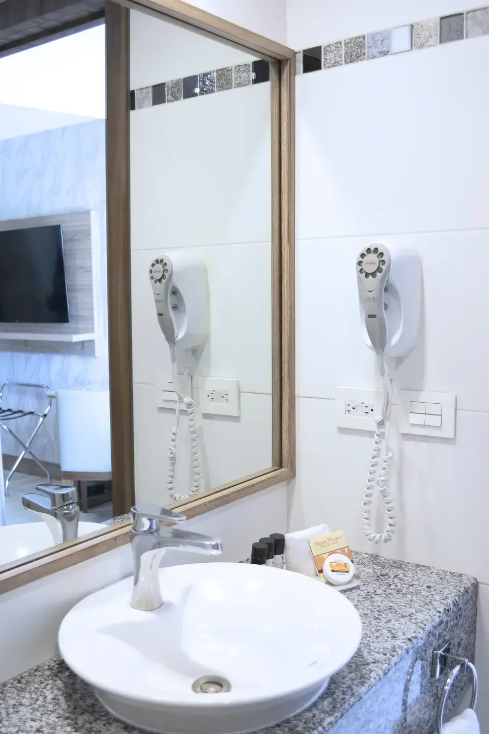 Lavabo pequeño con cubeta redonda blanca, espejo, secador de pelo montado en la pared y encimera de azulejos.