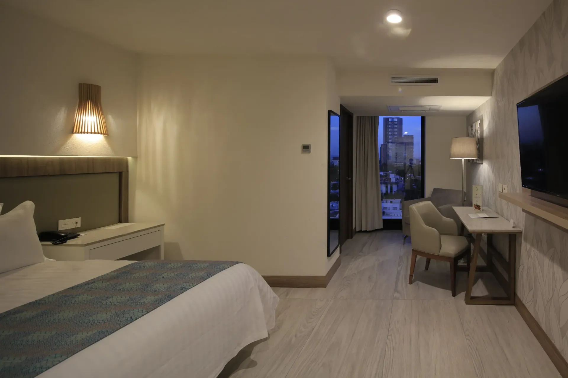 Habitación de hotel moderna con cama, escritorio, silla, lámpara, TV y balcón con vistas.