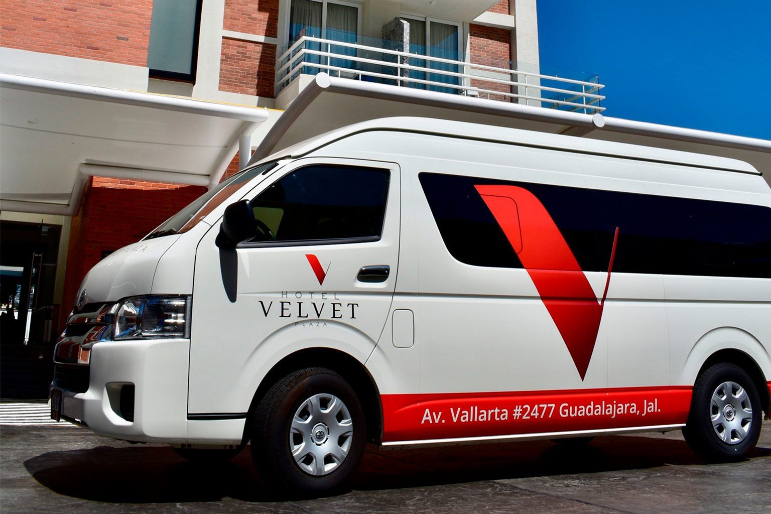 Furgoneta de pasajeros VELVET blanca con detalles rojos estacionada frente a un edificio.