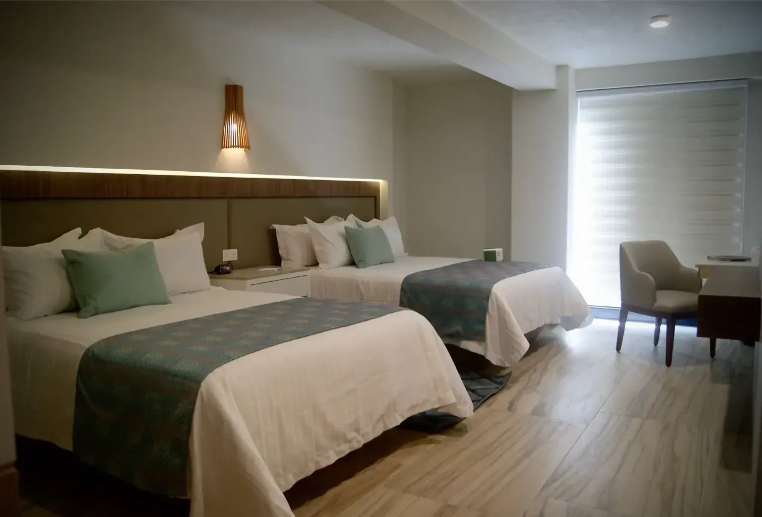 Habitación de hotel moderna con dos camas, detalles en verde suave, suelo de madera y zona de estar junto a la ventana.