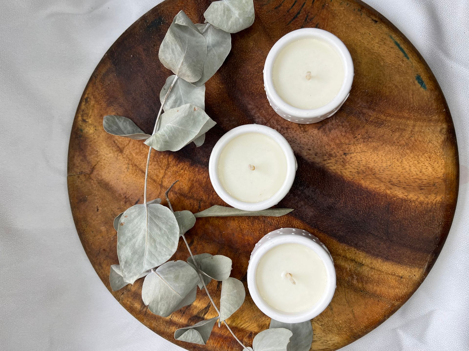 Tres velas blancas sobre una bandeja redonda de madera con hojas de eucalipto sobre un fondo gris.