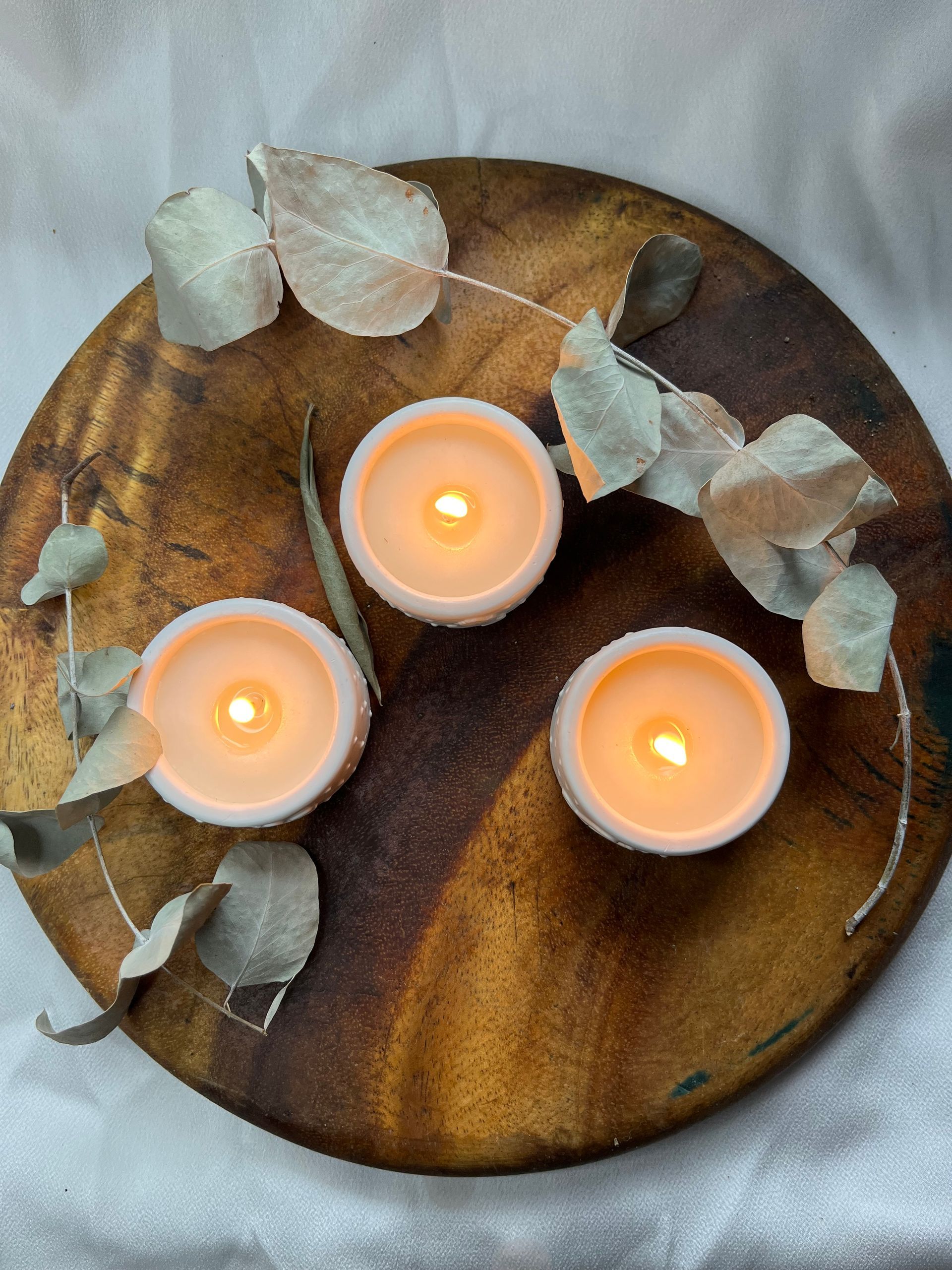 Tres velas blancas sobre una bandeja redonda de madera con hojas de eucalipto sobre un fondo gris.