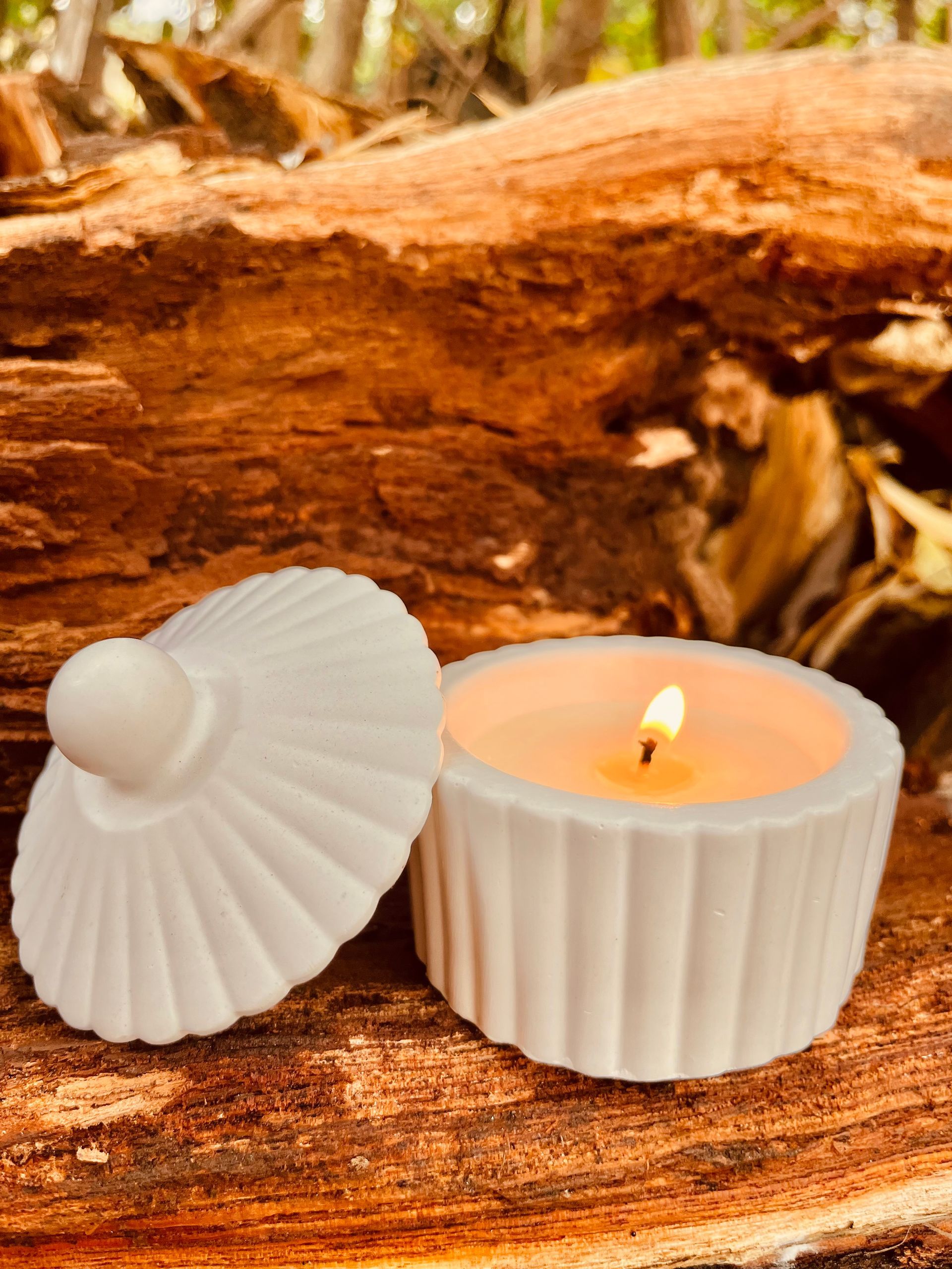 Vela blanca con forma de concha, con la llama encendida, junto a una pieza decorativa blanca a juego sobre madera rústica.