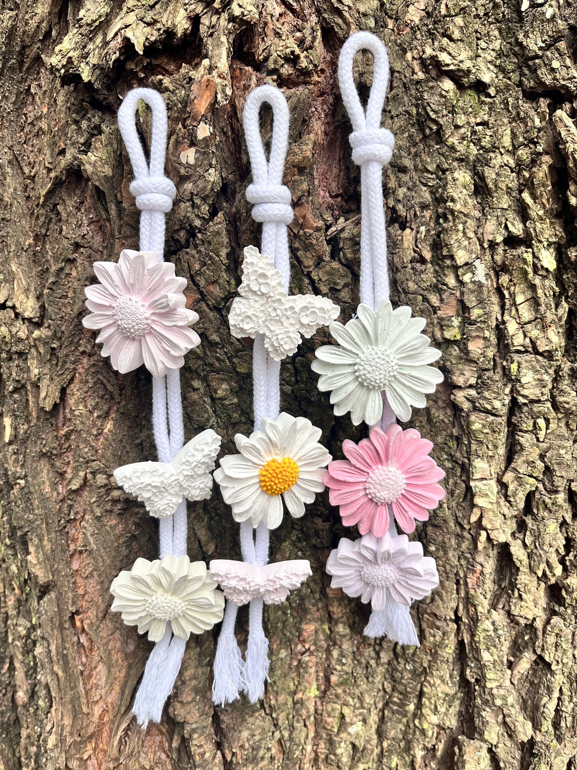 Tres lazos de cordón color lavanda con adornos de flores en tonos pastel dispuestos sobre corteza de árbol.