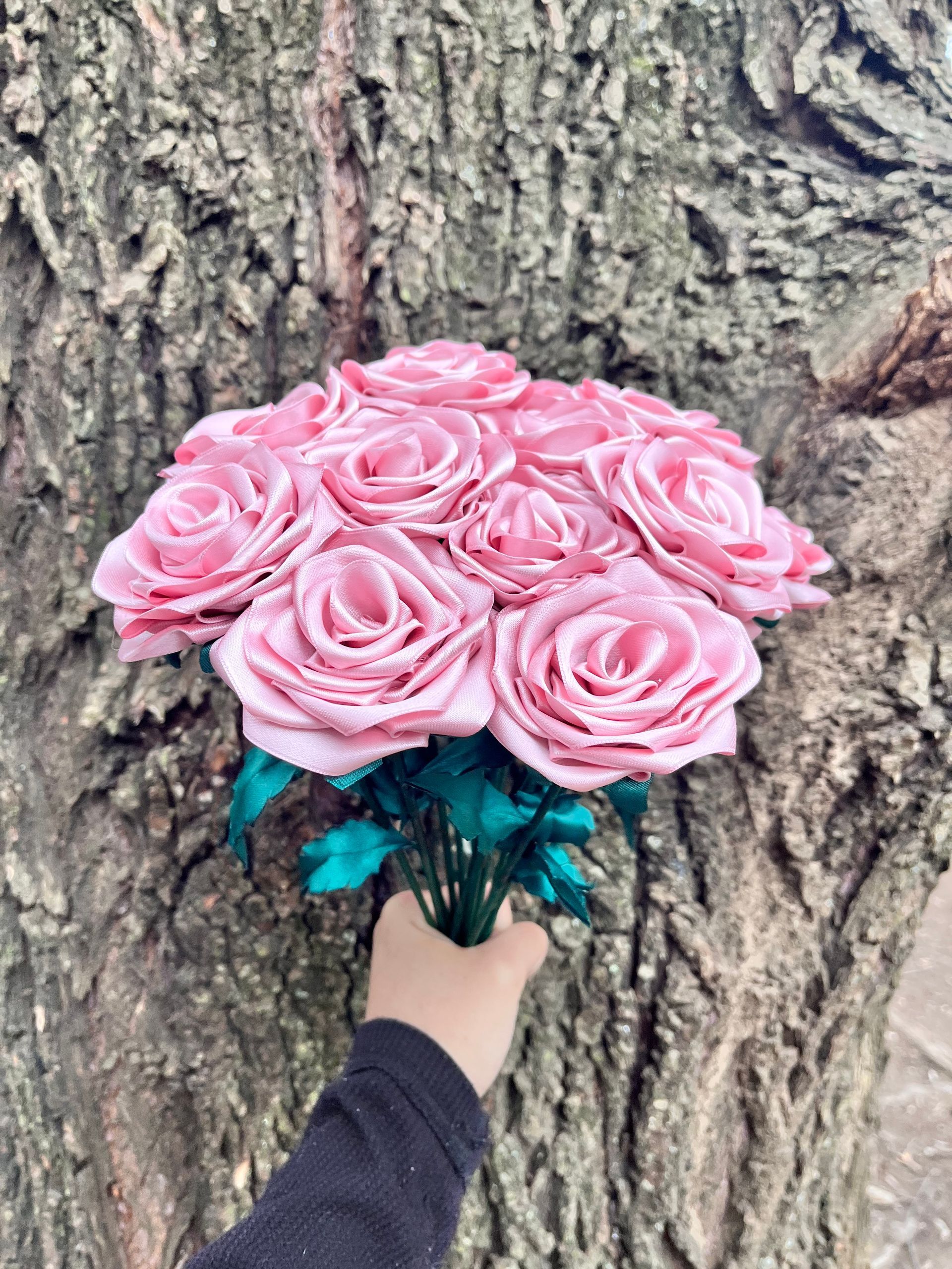 Una mano sostiene un ramo de rosas rosadas apoyado contra el tronco de un árbol con hojas turquesas.