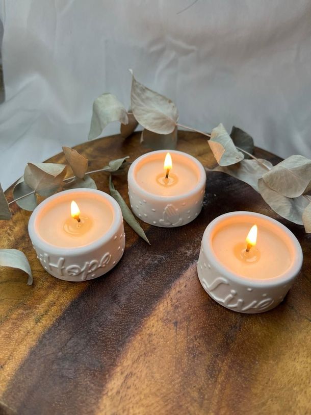 Tres velas pequeñas encendidas en portavelas blancos sobre una bandeja de madera con hojas decorativas.