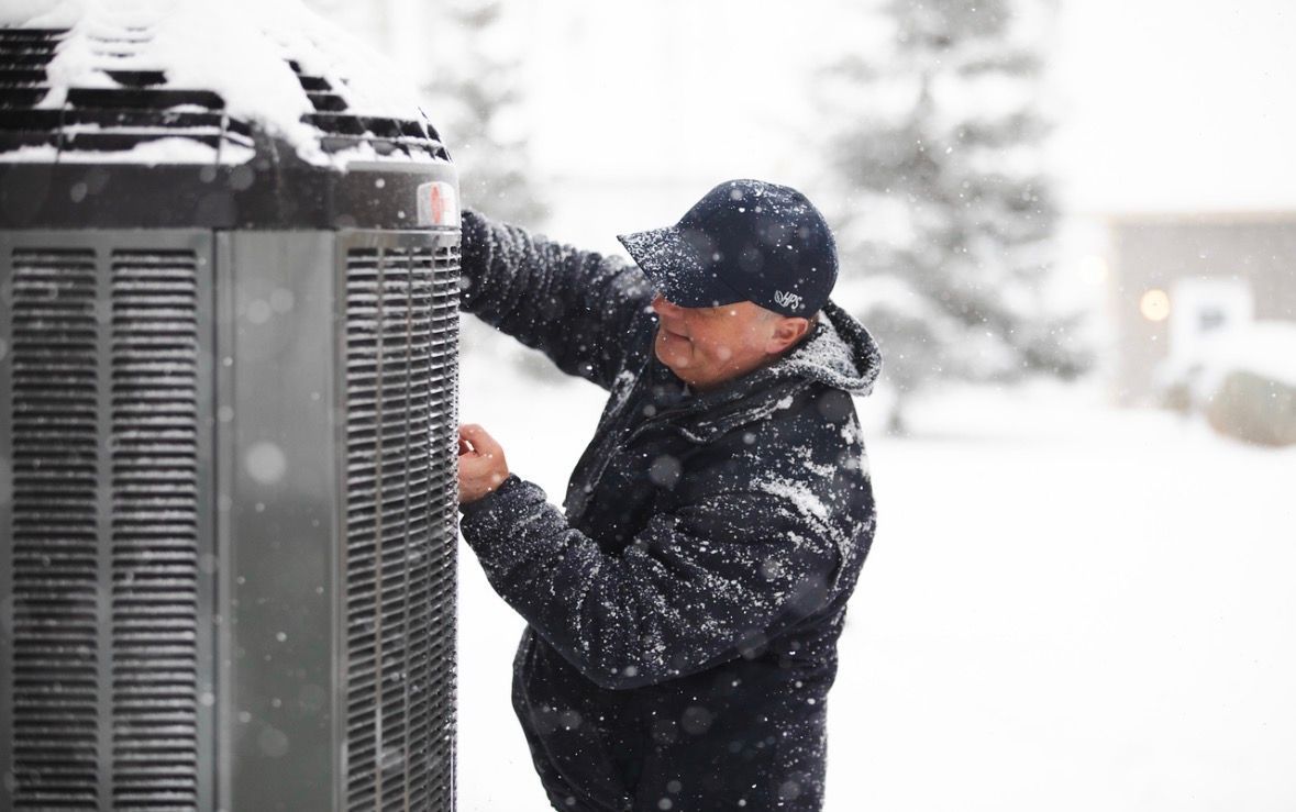 Man in dark coat and cap adjusting a large outdoor HVAC unit in heavy snowfall