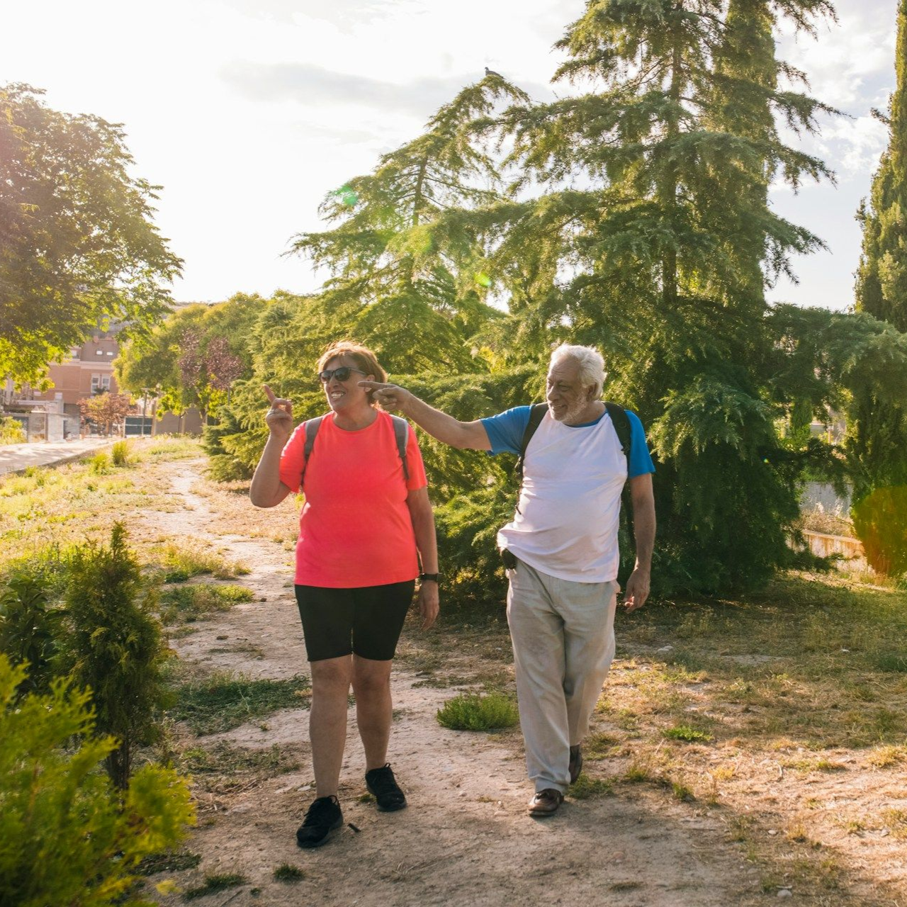 Elderly couple walking on a path through a sunny park. Man points, woman in pink, trees in background.