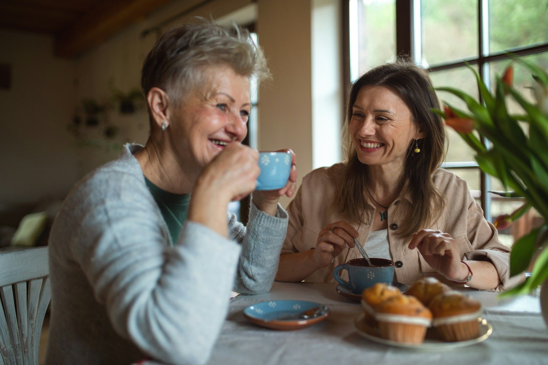 Two women smiling, having tea, muffins on a table near a window.