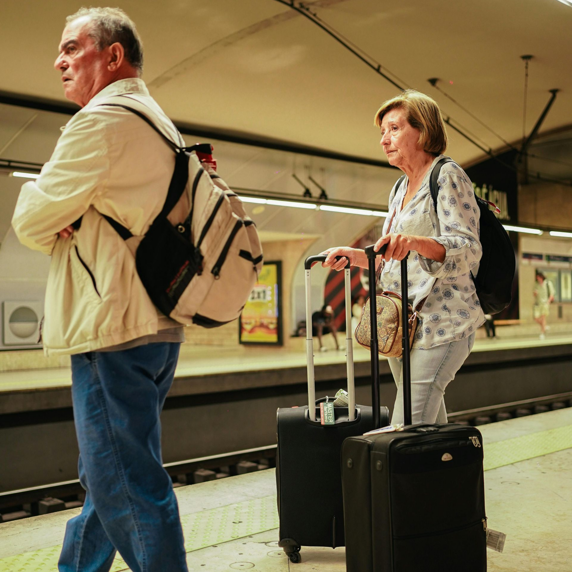 Two older adults with luggage at a subway platform, waiting for a train.