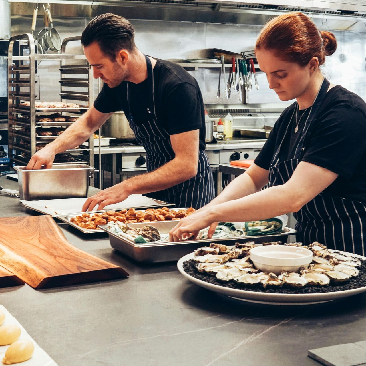 Chefs in black shirts and aprons plating food on a stainless steel counter in a kitchen.