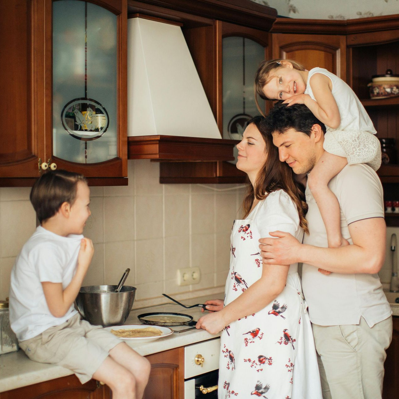Family in kitchen, making pancakes. Mother and father smiling, daughter on dad's shoulders, son looking on happily.