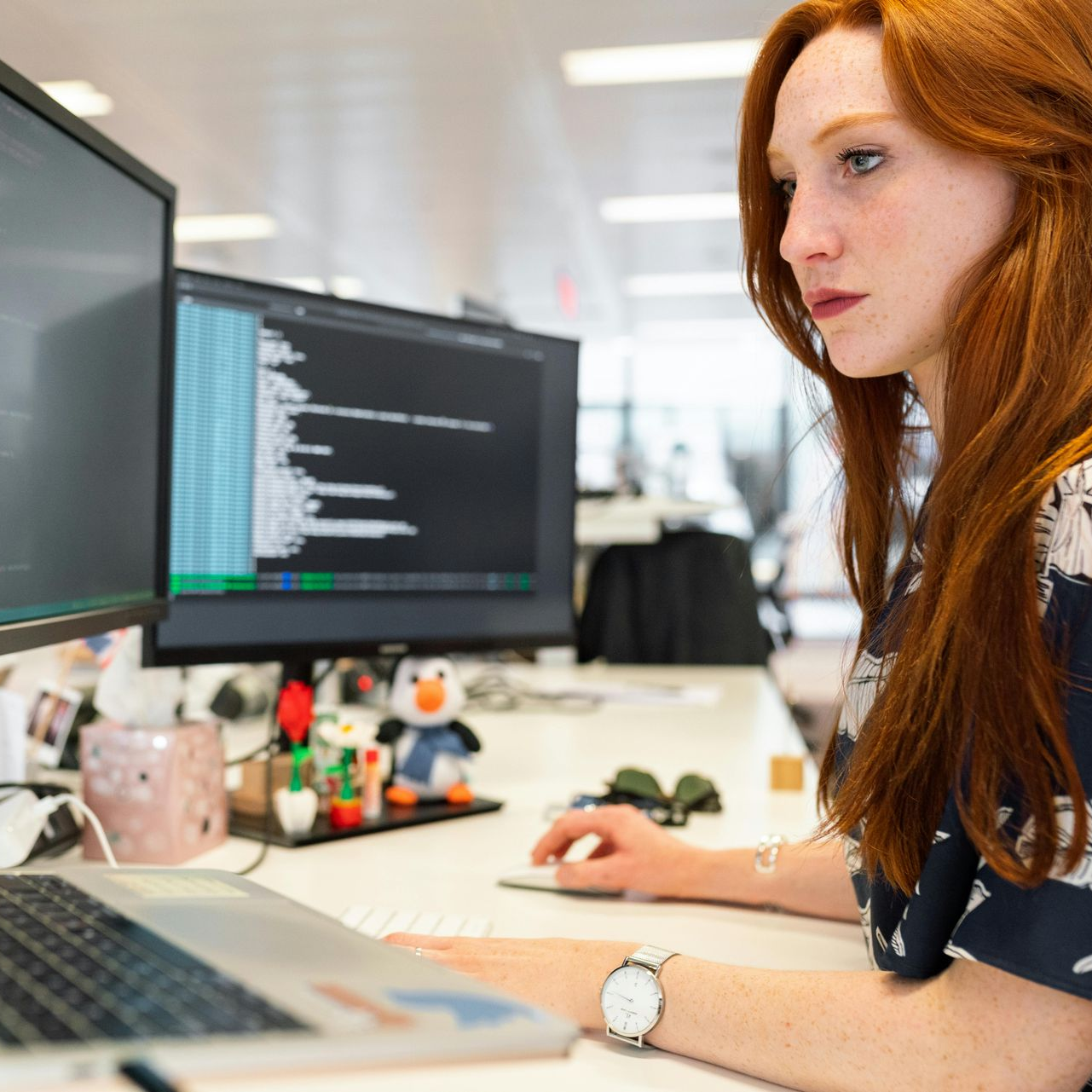 Woman with red hair coding on computer, focused expression, office setting.