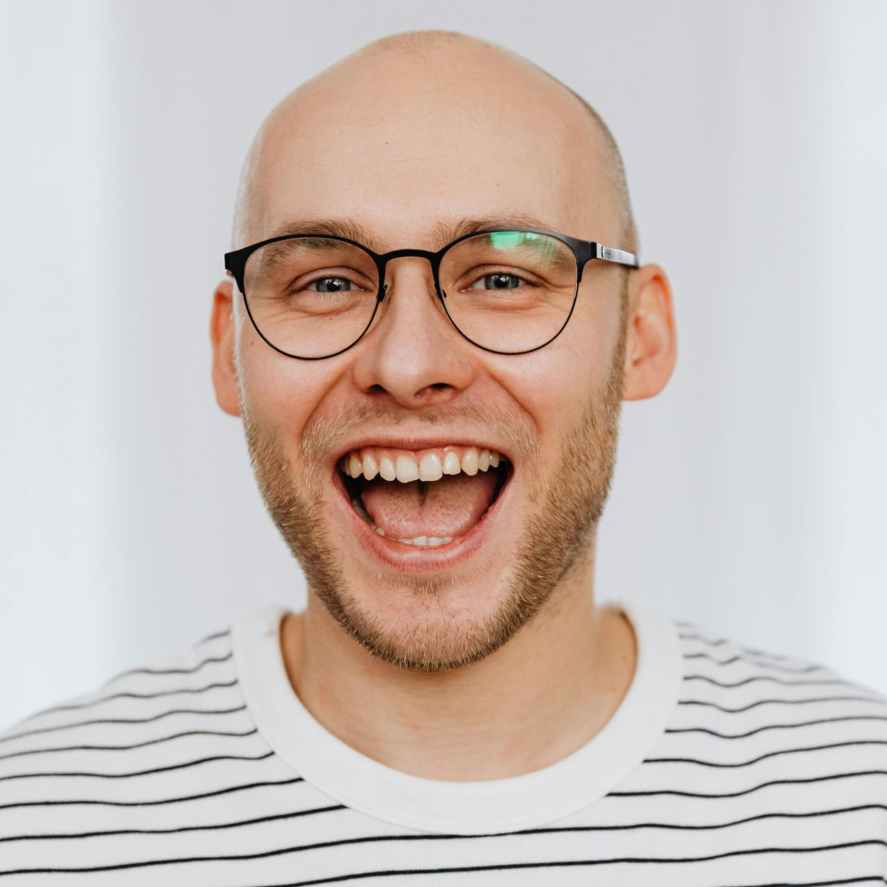 Bald man with glasses laughs, wearing a striped shirt, against a white background.