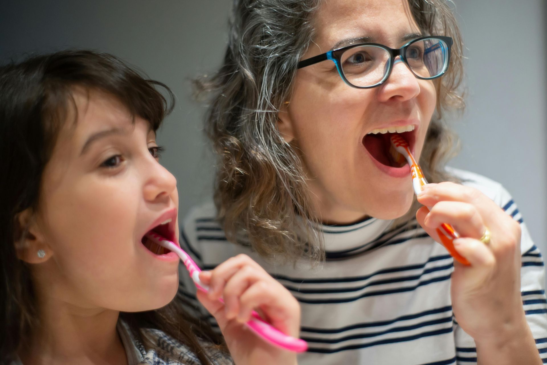 Woman and girl brushing their teeth together; bathroom setting.