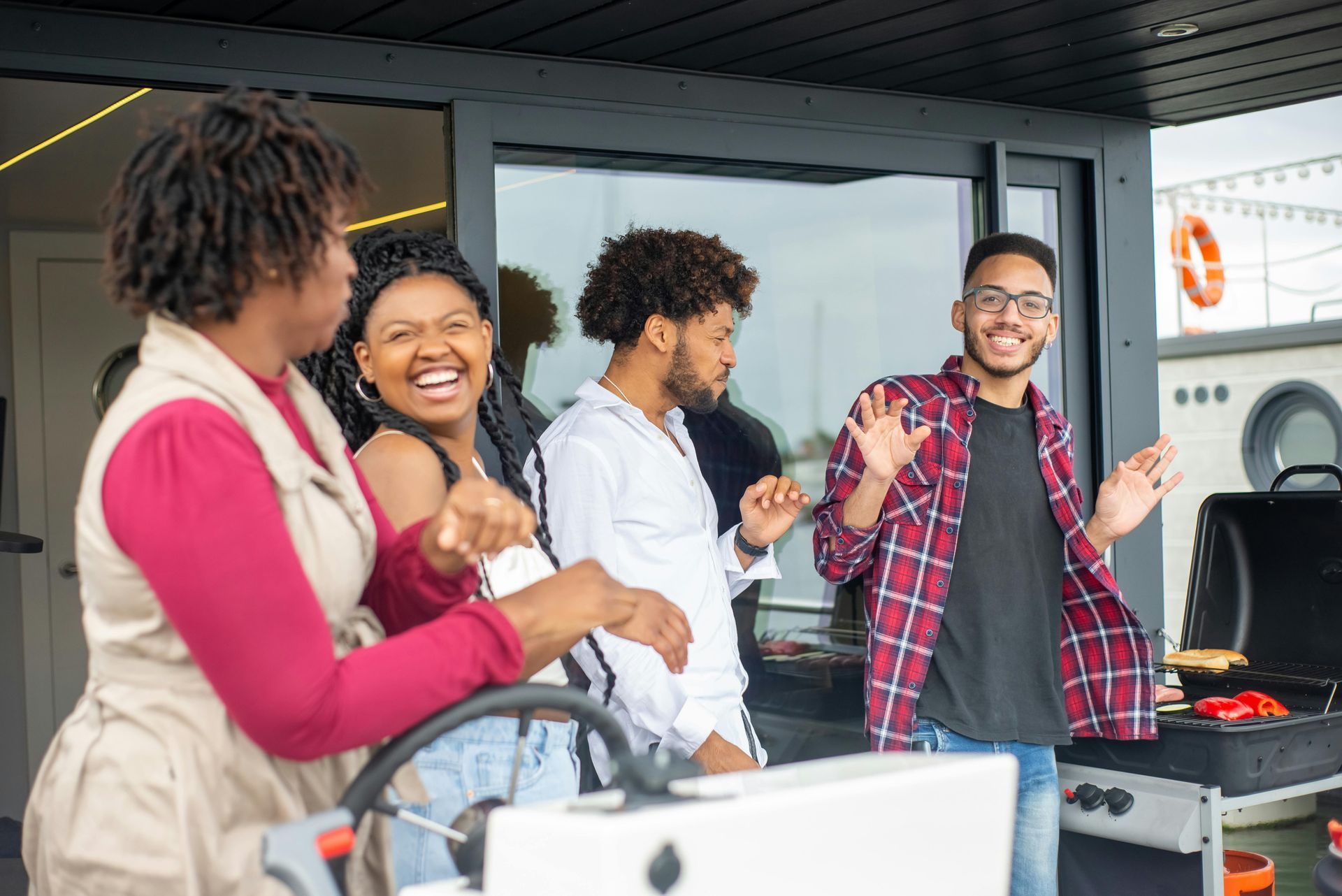 Group of friends laughing and grilling on a boat; person in red shirt waves.