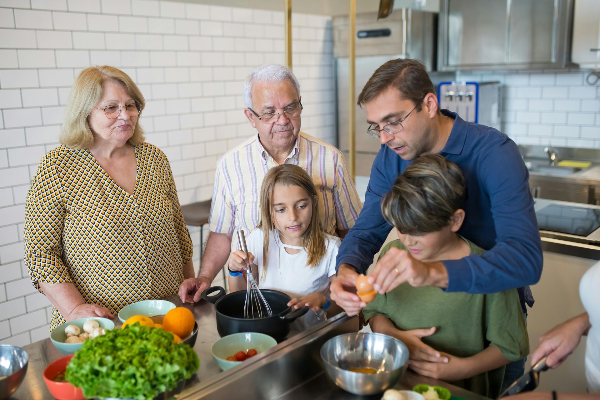 Family cooking together in a kitchen. Dad cracks an egg, kids mix, grandparents watch.