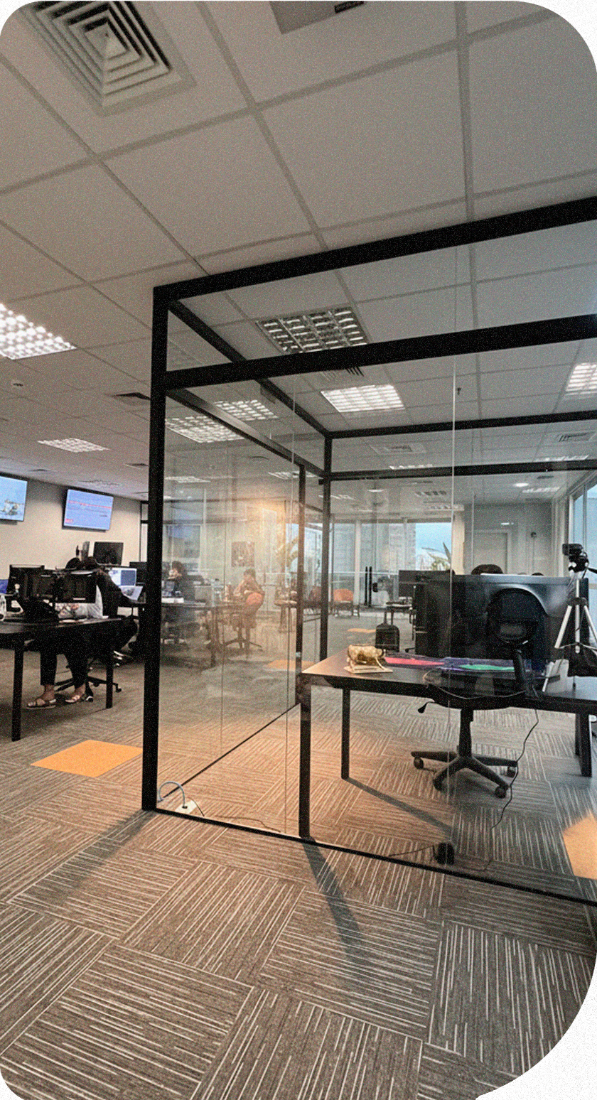 Espaço de trabalho interno com mesas, computadores e cadeiras. A luz do sol entra pelas janelas.