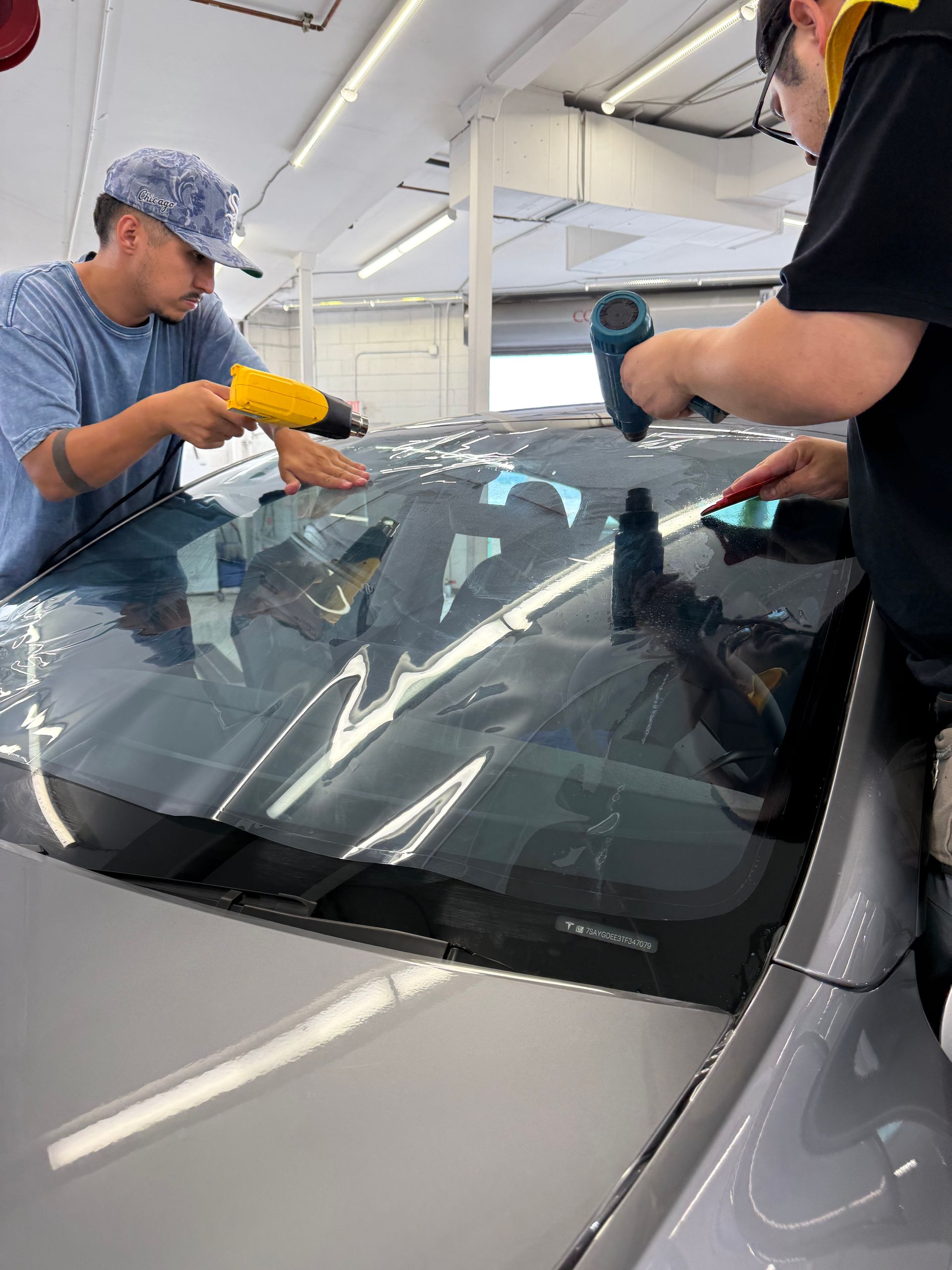 Two people applying window tint to a car window using heat guns in a workshop.