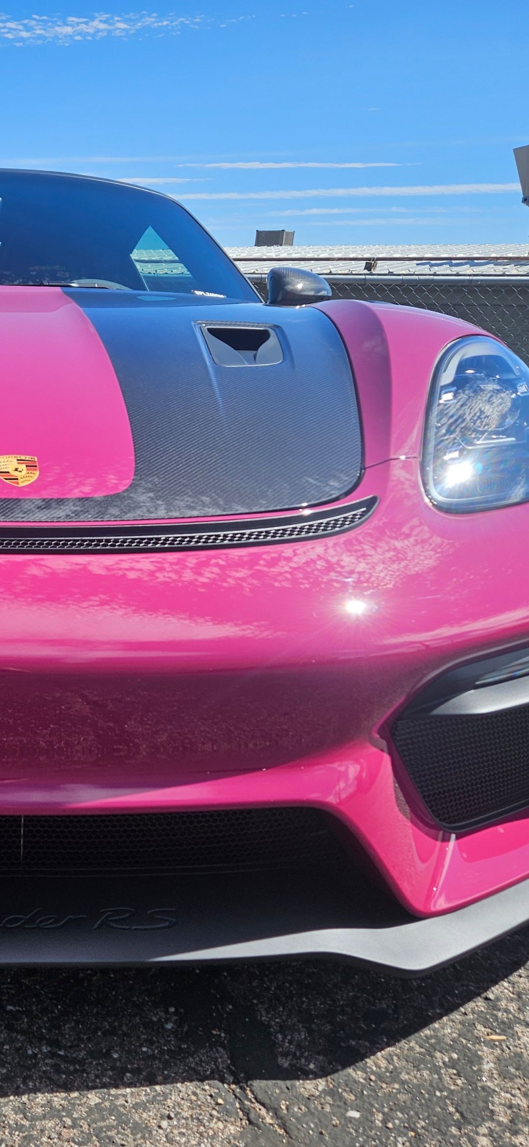 A bright pink sports car with a carbon fiber hood in an outdoor setting.