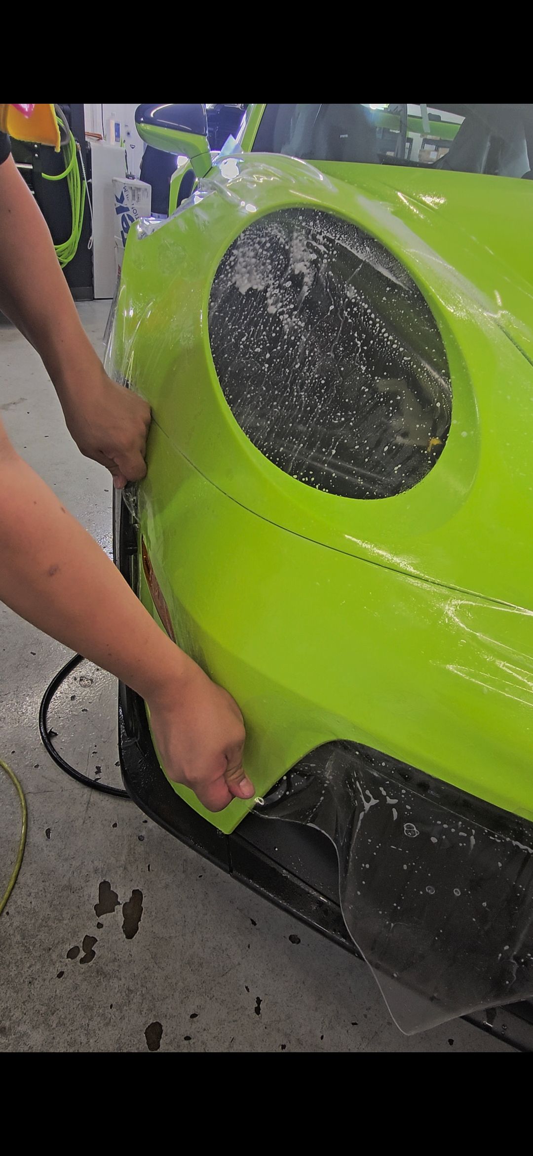 Person applying a protective film to the front of a neon green car bumper in a workshop.
