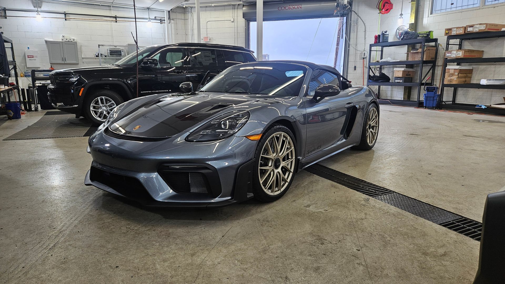 Gray Porsche sports car in a garage with a black pickup truck behind it.