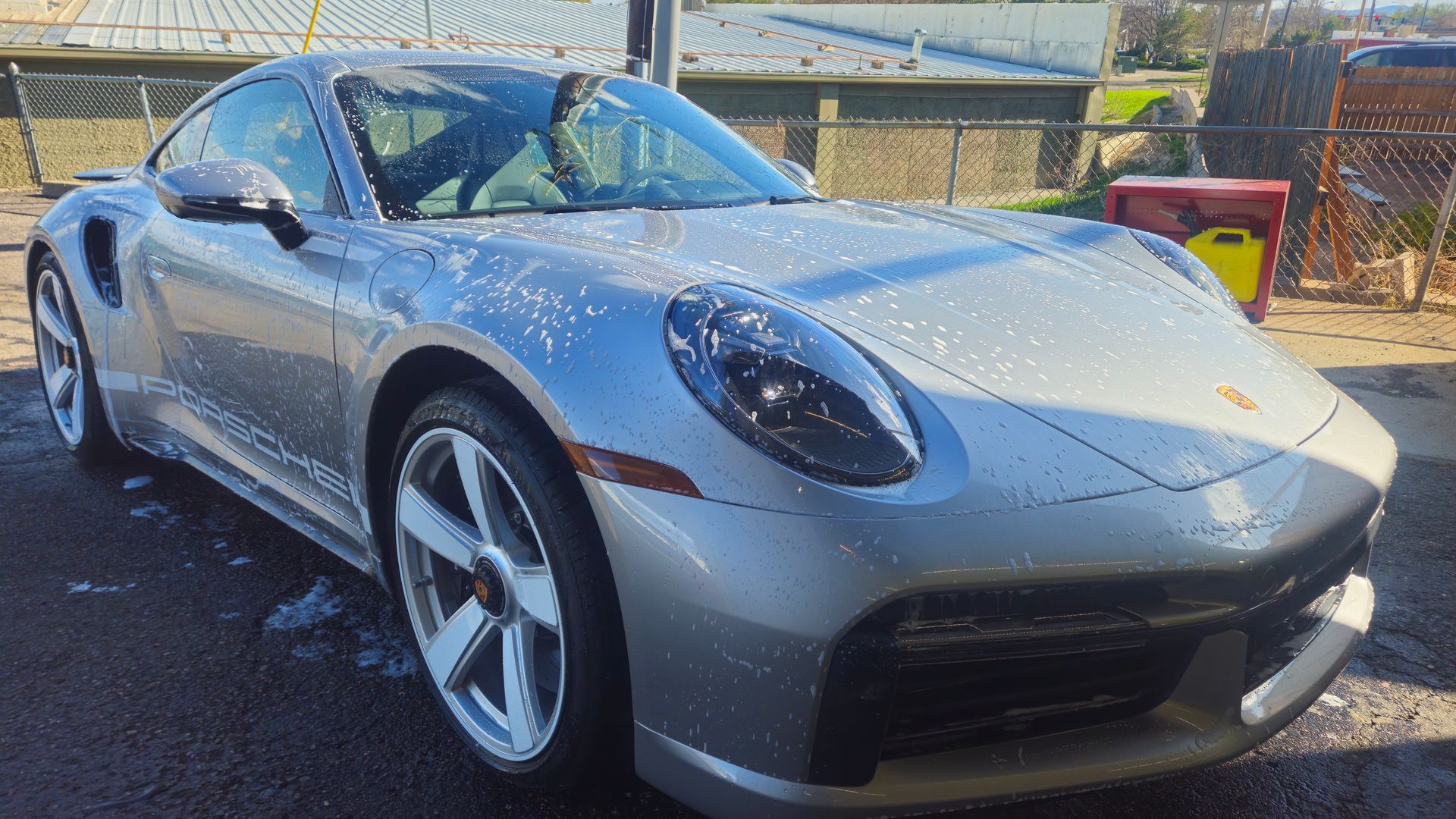 Silver Porsche 911 being washed at a car wash, covered in suds.