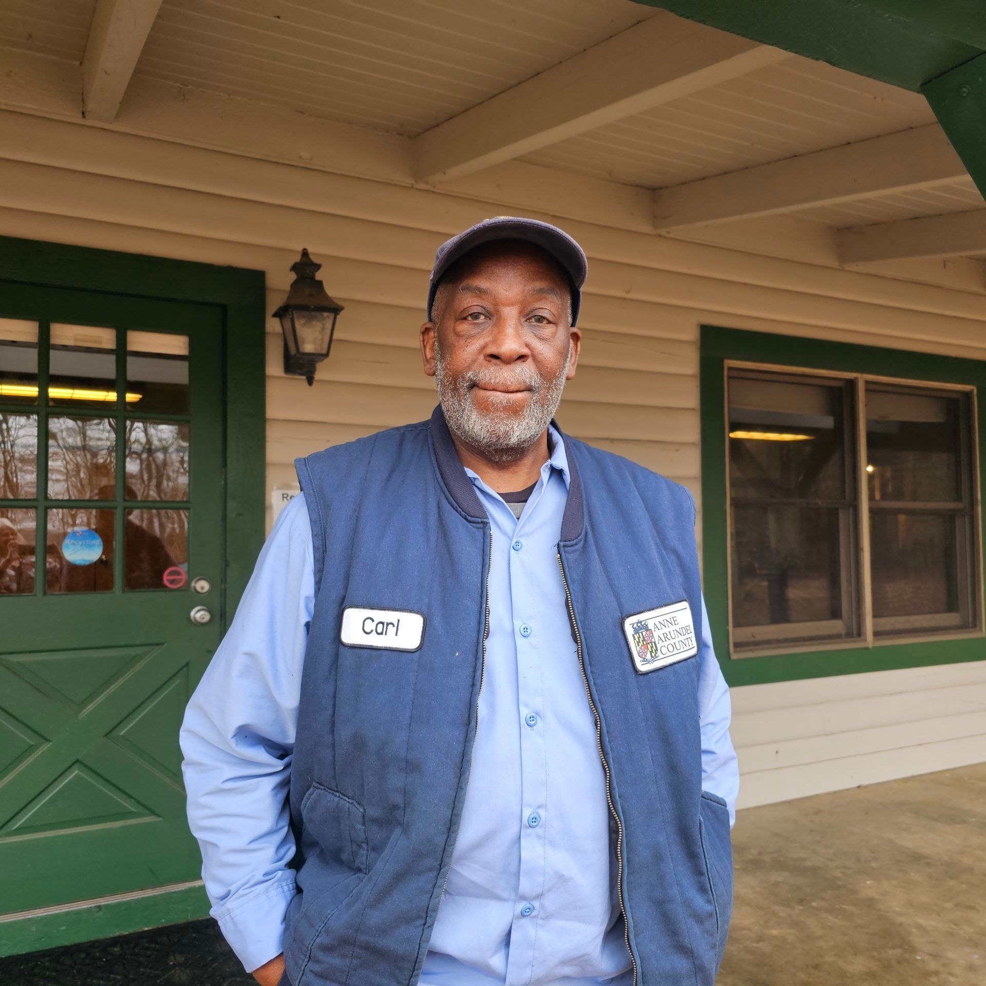 Man standing on a porch by a green door, wearing a blue shirt and vest with a name tag.
