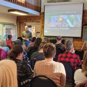 Bird presentation to seated audience in a cozy indoor hall with projector screen and slide titled “Prothonotary Warbler”