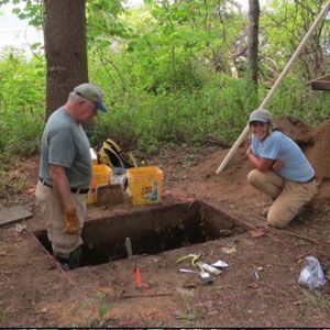 Two people excavating a rectangular pit in a wooded area beside a yellow machine