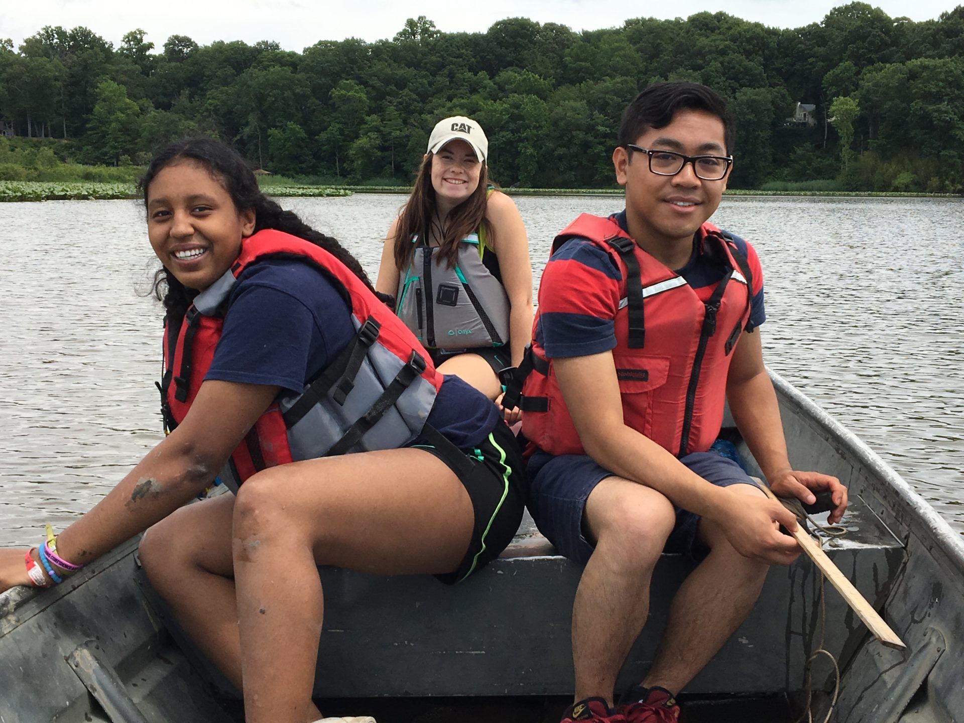Three friends in life jackets sitting in a small boat on a lake, smiling with trees in the background