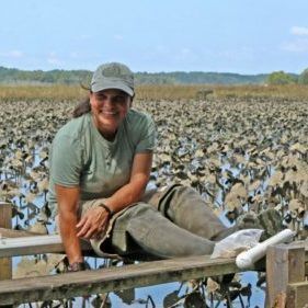 Person sitting on a wooden rail by a marsh, smiling in a green shirt and cap with birds filling the background