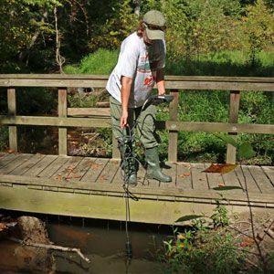 Person standing on a wooden bridge holding a rope over a small stream in a wooded area
