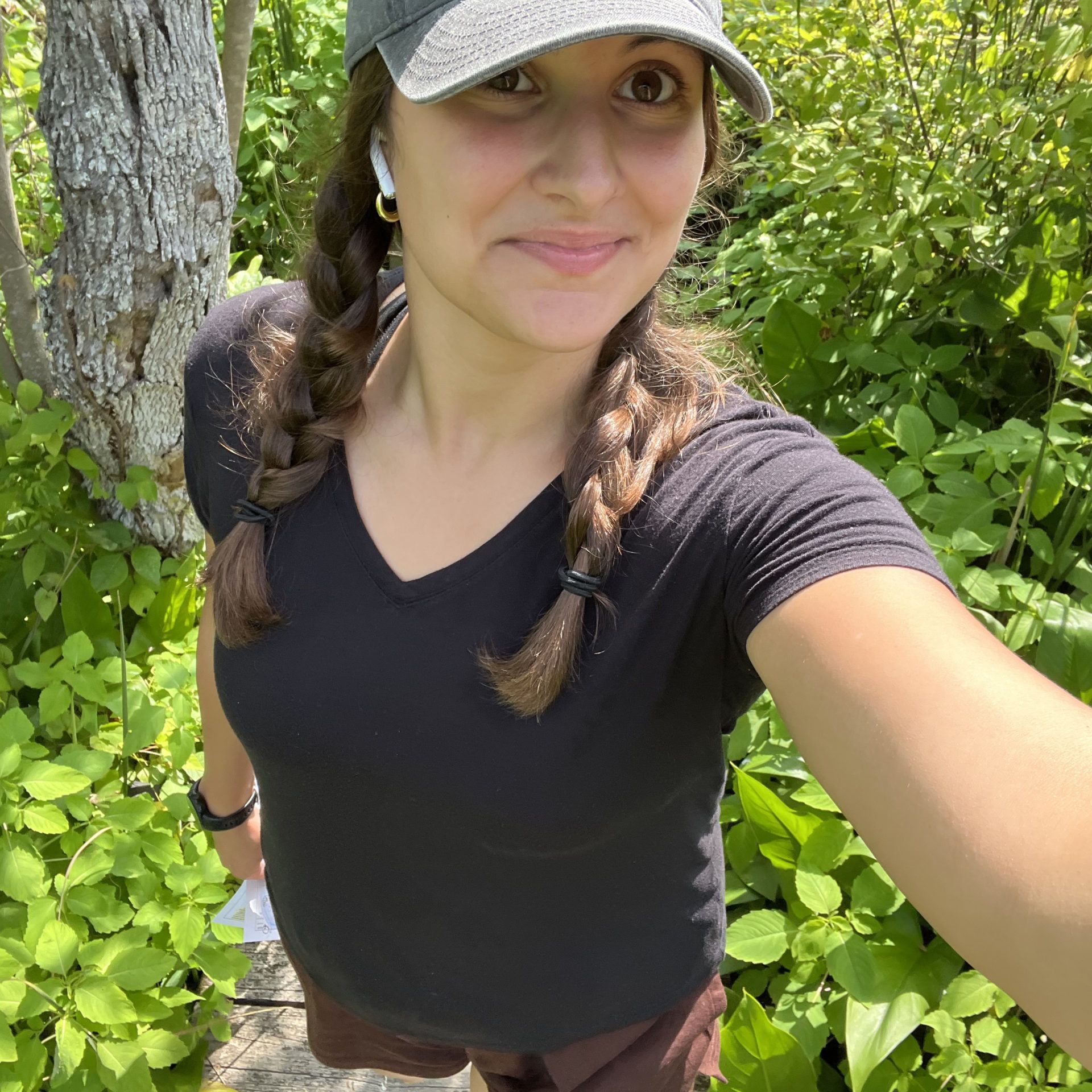 Woman in a black shirt and cap taking a selfie outdoors by a tree and green foliage
