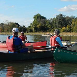 People in colorful canoes on a calm river, with trees and blue sky in the background