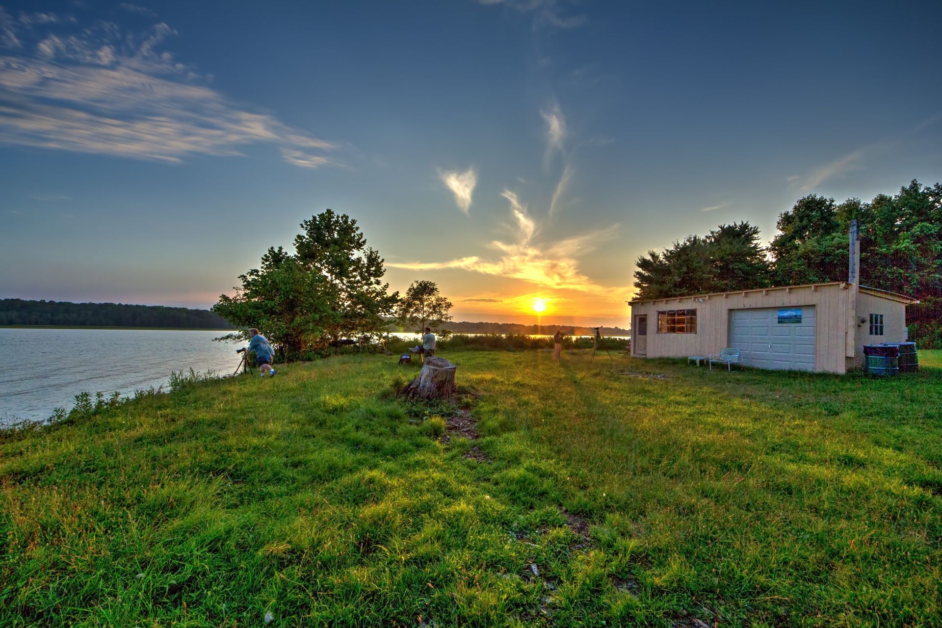 Sunset over a grassy lakeside with a small white building and trees under a colorful sky