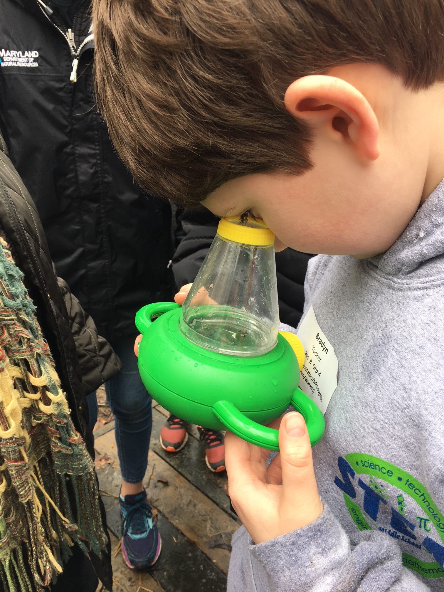 Child peers into a clear container held in a bright green holder during an outdoor activity
