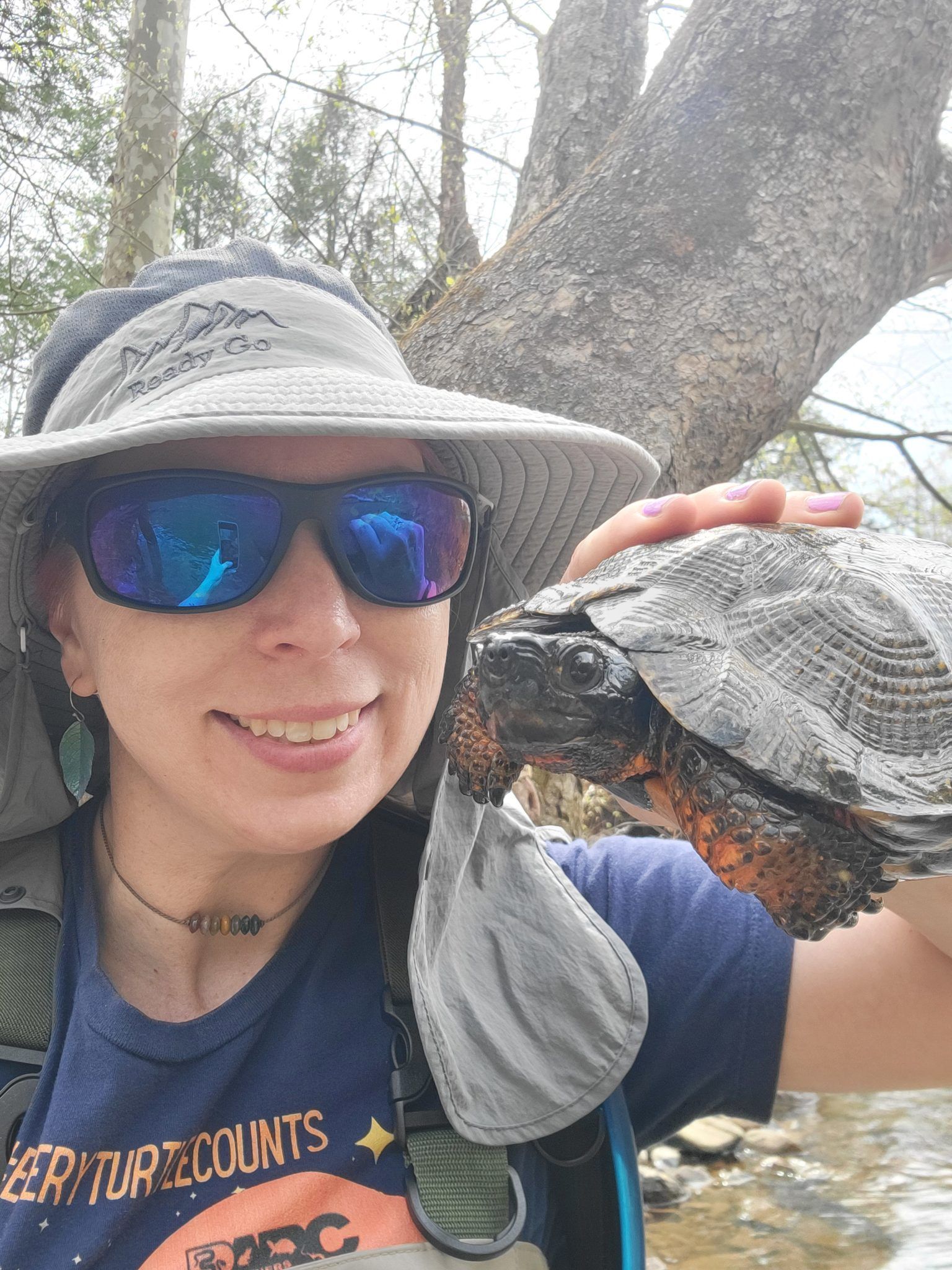 Hiker in sun hat and sunglasses holding a shell by a creek under trees