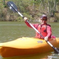 Person in a red jacket and cap kayaking on a river in a yellow kayak, holding a paddle.