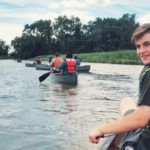 Two people canoeing on a river, with a smiling person in a red life jacket in the foreground.